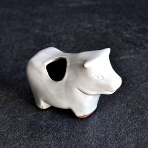 May include: A white ceramic bull figurine with a large opening on its side. The sculpture has a smooth finish and a small brown base. The bull is depicted in a standing position, with its head turned slightly to the side.