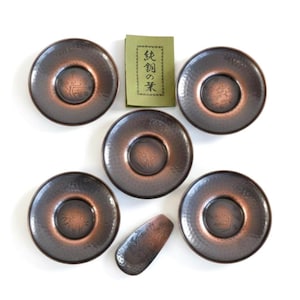 Può includere: Set di sei piattini da tè color rame con texture martellata e design centrale. Include un cucchiaino abbinato e un pacchetto di carta verde con testo giapponese. I piattini sono disposti su una superficie bianca.