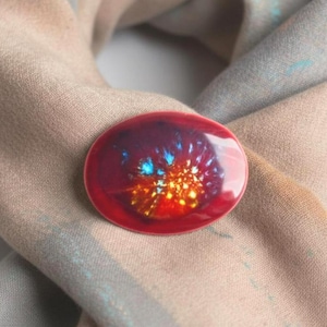 Peut inclure: Broche ovale rouge avec un motif coloré et vibrant. La broche présente un motif central aux teintes orange, bleu et jaune, sur un fond rouge foncé. La broche est posée sur une écharpe beige et grise.