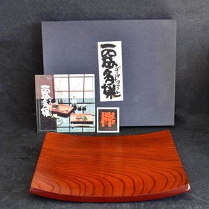 Peut inclure: Plateau rectangulaire en bois brun rougeâtre avec un bord incurvé et un motif de grain de bois visible. L'image présente également trois petites cartes et une boîte bleu foncé en arrière-plan, avec du texte japonais sur les cartes.