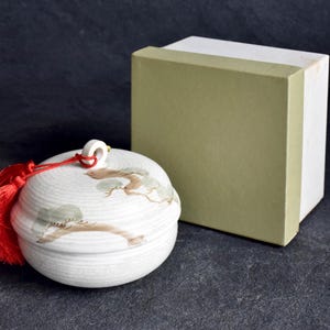 Könnte beinhalten: Ein weißer Keramikbehälter mit Deckel, bemaltem Baumdesign und roter Quaste. Neben dem Behälter befindet sich eine rechteckige Box mit grünem Äußeren und weißem Innenraum. Der Hintergrund ist dunkel.