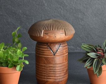 May include: A hand-carved wooden figurine featuring a smooth, rounded head with etched hair lines and a cylindrical body with horizontal grooves. The figure has a V-shaped neckline carving and closed eyes, presented in a natural brown wood grain finish.