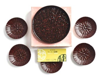 May include: A set of five dark brown, circular, woven-textured lacquerware plates arranged around a central square box. A yellow rectangular label with Japanese text and bamboo illustrations is placed between the plates and the box.