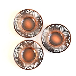 May include: Three decorative copper-coloured plates with intricate openwork designs. Each plate features a central, slightly recessed area and a detailed border with a pattern of small white dots and floral accents. The plates are arranged on a white surface.
