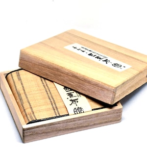 May include: A wooden box with a lid, containing a folded textile. The textile is beige with vertical black stripes. The box has a label on the lid with Japanese text. The box is open, revealing the contents.