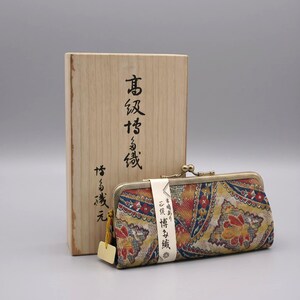 May include: A rectangular fabric coin purse with a gold-toned metal kiss-lock clasp, featuring a multi-colored floral and geometric pattern. A white paper band with Japanese text wraps around the purse, which sits before a wooden box.