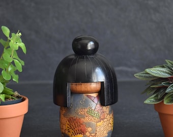 May include: A wooden kokeshi doll featuring a smooth, black, rounded hairstyle and a natural wood-toned face. The cylindrical body is decorated with a detailed, multi-colored pattern in shades of blue, orange, and yellow, resembling a kimono.