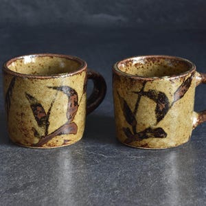 Może przedstawiać: Dwa ceramiczne kubki o rustykalnym, ręcznie robionym wyglądzie. Kubki mają beżowy kolor bazowy z ciemnobrązowymi wzorami w kształcie liści. Obręcze i uchwyty są ciemnobrązowe. Kubki są prezentowane na ciemnoszarej powierzchni.