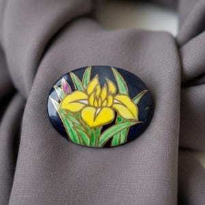Könnte beinhalten: Eine ovale Brosche mit dunkelblauem Hintergrund, verziert mit einer handgemalten gelben Irisblüte und grünen Blättern. Das florale Design ist mit feinen goldfarbenen Metalllinien umrandet, die einen deutlichen Kontrast zur tiefblauen Emaille bilden.