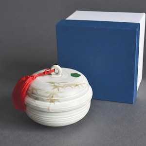 Puede incluir: Un recipiente de cerámica blanca con tapa, con una borla roja y un acento de hoja verde. El recipiente tiene un diseño de hojas de bambú y está junto a una caja azul y blanca. Es un objeto decorativo.