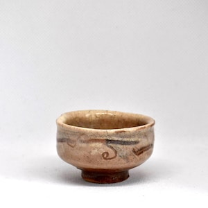 Può includere: Una piccola ciotola da tè in ceramica rotonda con una finitura smaltata color beige chiaro crepata. L'esterno presenta motivi decorativi lineari e a spirale dipinti a mano in marrone scuro. La ciotola ha un piede spesso e un bordo irregolare.