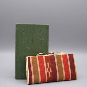 May include: A rectangular clutch bag featuring a woven fabric with vertical stripes in shades of red, burgundy, and tan, finished with a gold-tone metal frame and a kiss-lock clasp, accompanied by a tall, textured dark green rectangular box.