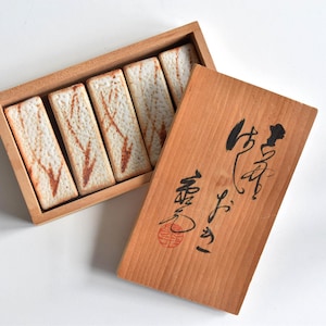 May include: A rectangular wooden box containing five rectangular cookies with a light brown marbled pattern. The wooden lid is placed beside the box, featuring black Japanese calligraphy and a small red circular stamp on the surface.