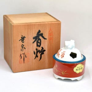 May include: A Japanese incense burner with a red, white, and blue design, and a wooden box. The burner has a round shape with a lid and a base. The wooden box has Japanese characters on it.
