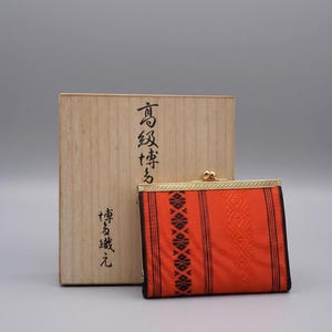 Op de afbeelding: Een rechthoekige oranje portemonnee met zwarte verticale strepen en een geometrisch patroon, voorzien van een goudkleurige metalen knipsluiting. Het staat voor een lichtgekleurde houten doos met zwarte Japanse kalligrafie.