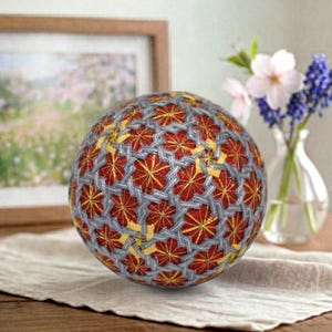 Puede incluir: Una bola Temari artesanal con un complejo diseño geométrico. La bola está hecha con hilo rojo, gris y amarillo. Descansa sobre una tela blanca, con una imagen enmarcada y un jarrón de flores en el fondo.