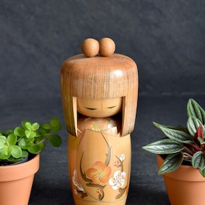 Könnte beinhalten: Eine Kokeshi-Puppe aus hellem Holz mit einem abgerundeten Kopf, auf dem sich zwei kleine Holzkugeln befinden. Der Körper der Puppe ist mit floralen Designs in Orange und Weiß verziert. Zwei kleine Topfpflanzen flankieren die Puppe.