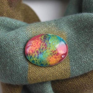 May include: An oval brooch with a vibrant, abstract pattern in pink, orange, yellow, green, and blue. The brooch is pinned to a green and brown plaid scarf.