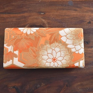 Puede incluir: Un estuche rectangular cubierto de tela naranja con un patrón tejido de grandes flores de crisantemo blancas y doradas y formas geométricas. La superficie tiene una apariencia texturizada con un motivo floral repetitivo.