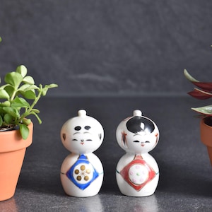 May include: Two small, white ceramic kokeshi-style figurines with rounded heads and bodies. The left figurine features a blue diamond pattern with a gold floral design. The right figurine has a black painted hairstyle and a red diamond pattern with dots.
