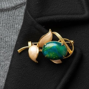 Könnte beinhalten: Eine Brosche aus goldfarbenem Metall mit einem zentralen, ovalen Cabochon-Stein mit einer gesprenkelten grünen und blauen Oberfläche. Die Brosche ist mit zwei glatten, cremefarbenen Emaille-Blättern und einem strukturierten, geschwungenen Stiel verziert.