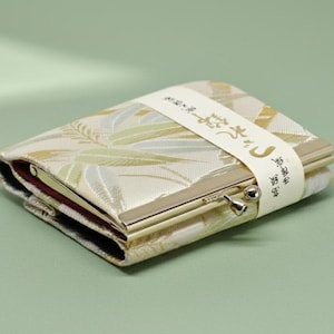 Può includere: Un piccolo portafoglio rettangolare con una chiusura in metallo color oro. Il portafoglio è avvolto da una fascia bianca con testo giapponese. Il tessuto ha uno sfondo color crema con un motivo a foglie in oro e verde chiaro.