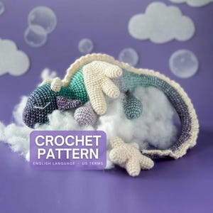 May include: A crocheted dragon in shades of blue, green, and cream, resting on a cloud of fluffy material. The dragon is accompanied by small crocheted spheres and a star. The image includes the text "CROCHET PATTERN" and "ENGLISH LANGUAGE • US TERMS".