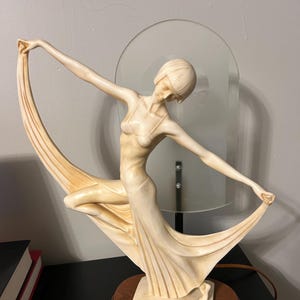 Könnte beinhalten: Elfenbeinfarbene Art-Deco-Lampe mit einer stilisierten weiblichen Figur, die in fließendem Stoff drapiert ist. Die Figur ist mit ausgestreckten Armen dargestellt, die den Stoff halten. Die Lampe steht auf einem Holzsockel mit runder Form. Hinter der Figur befindet sich eine Milchglasscheibe.