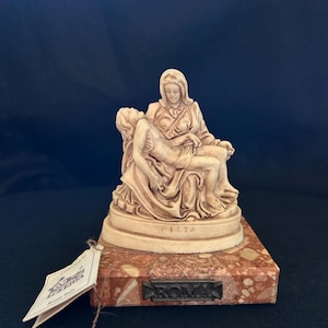 Könnte beinhalten: Beigefarbene Skulptur, die die Pietà darstellt, mit der Jungfrau Maria, die den Körper Jesu hält. Die Skulptur ist auf einem rechteckigen, gesprenkelten, rotbraunen Sockel montiert. Das Wort "PIETA" ist auf der Skulptur und "ROMA" auf dem Sockel eingraviert.