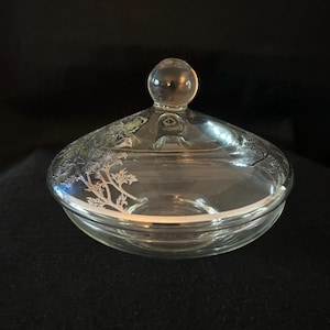 Op de afbeelding: Een heldere glazen bonbonnière met een deksel. Het deksel heeft een ronde knop en een zilveren bloemenontwerp rond de rand. De schaal staat op een zwart oppervlak.