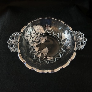 Op de afbeelding: Een heldere glazen kom met een zilveren rand en bloemenmotief. De kom heeft twee sierlijke handvatten en een geschulpte rand. Het bloemenpatroon is in het glas geëtst, wat een delicate en elegante uitstraling creëert.