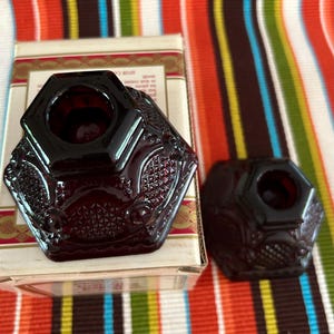 May include: Two ruby red glass candle holders with ornate, geometric designs. One is on a box with text, the other is separate. The candle holders have a hexagonal shape with a central circular opening.