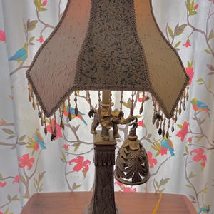 Puede incluir: Una lámpara de mesa ornamentada con una pantalla estampada en tonos beige y marrón. La lámpara presenta una base decorativa con figuras de elefantes y un adorno en forma de campana. La pantalla está adornada con flecos de cuentas.