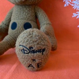 May include: A brown plush toy with a Disney logo embroidered on the foot. The toy has a fuzzy texture, two black circular accents, and a light orange background.