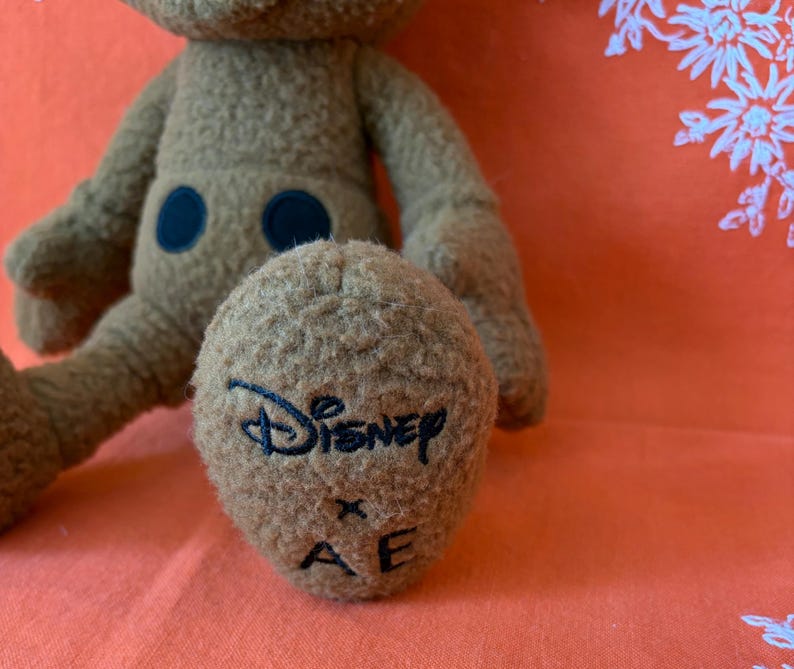 May include: A brown plush toy with a Disney logo embroidered on the foot. The toy has a fuzzy texture, two black circular accents, and a light orange background.
