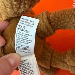 May include: A close-up of a brown plush toy with a tag attached. The tag includes washing instructions and the text "AMERICAN EAGLE OUTFITTERS". The toy is made of polyester, plastic, and polyurethane.