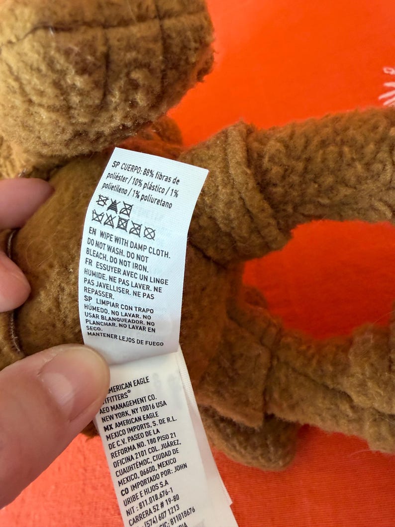 May include: A close-up of a brown plush toy with a tag attached. The tag includes washing instructions and the text "AMERICAN EAGLE OUTFITTERS". The toy is made of polyester, plastic, and polyurethane.