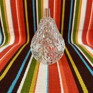 May include: A clear, faceted glass perfume bottle with a silver stopper. The bottle has a teardrop shape and sits on a colourful striped background. The glass is cut with a diamond pattern, reflecting light.