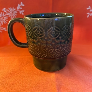 Può includere: Tazza in ceramica verde scuro con manico. La tazza presenta un motivo in rilievo ripetuto di disegni floreali stilizzati. La tazza è posta su uno sfondo arancione.