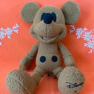 May include: A brown plush Mickey Mouse toy with large ears, a black nose, and two black button details on the front. The Disney logo is embroidered on one foot. The toy is sitting on an orange surface.