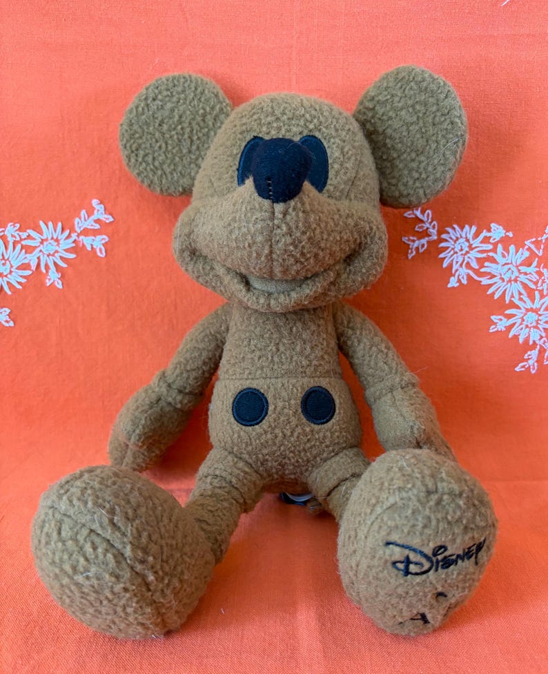 May include: A brown plush Mickey Mouse toy with large ears, a black nose, and two black button details on the front. The Disney logo is embroidered on one foot. The toy is sitting on an orange surface.