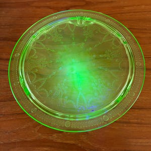 May include: A round, bright green glass tray with etched floral and geometric patterns. The tray has a decorative border and a central design. The glass has a slight glow, and the tray is set on a wooden surface.