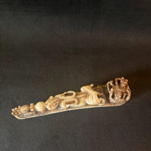 Può includere: Scultura scolpita color avorio con figure dettagliate di animali e persone. Il pezzo allungato presenta una serie di elementi scolpiti, tra cui una sfera centrale, figure di animali e una figura coronata all'estremità. Sfondo scuro.