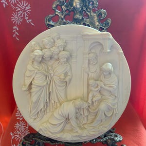 May include: Off-white circular relief sculpture depicting a religious scene with multiple figures. The sculpture is mounted on a decorative metal stand. The figures are dressed in draped garments, and the scene appears to be a biblical narrative.