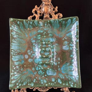 Può includere: Un piatto decorativo quadrato con una glassa screziata verde acqua e marrone, posto su un supporto in metallo dorato. Il piatto ha angoli arrotondati e una superficie strutturata. Il supporto presenta dettagli ornati.