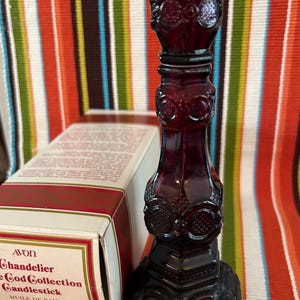Op de afbeelding: Een donkerrode glazen kandelaar met sierlijke details, onderdeel van de Avon God Collection. De basis is zeshoekig en de body heeft herhaalde ovale ontwerpen. Een doos met de tekst "AVON Chandelier God Collection Candlestick" is gedeeltelijk zichtbaar.