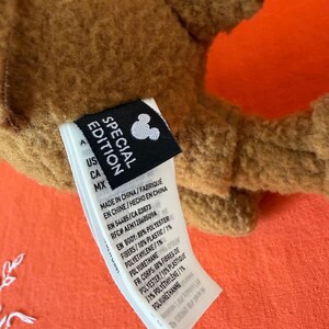 May include: Close-up of a brown plush toy with an orange accent. A black tag with the text "SPECIAL EDITION" and a Mickey Mouse silhouette is attached. The tag also includes manufacturing details.
