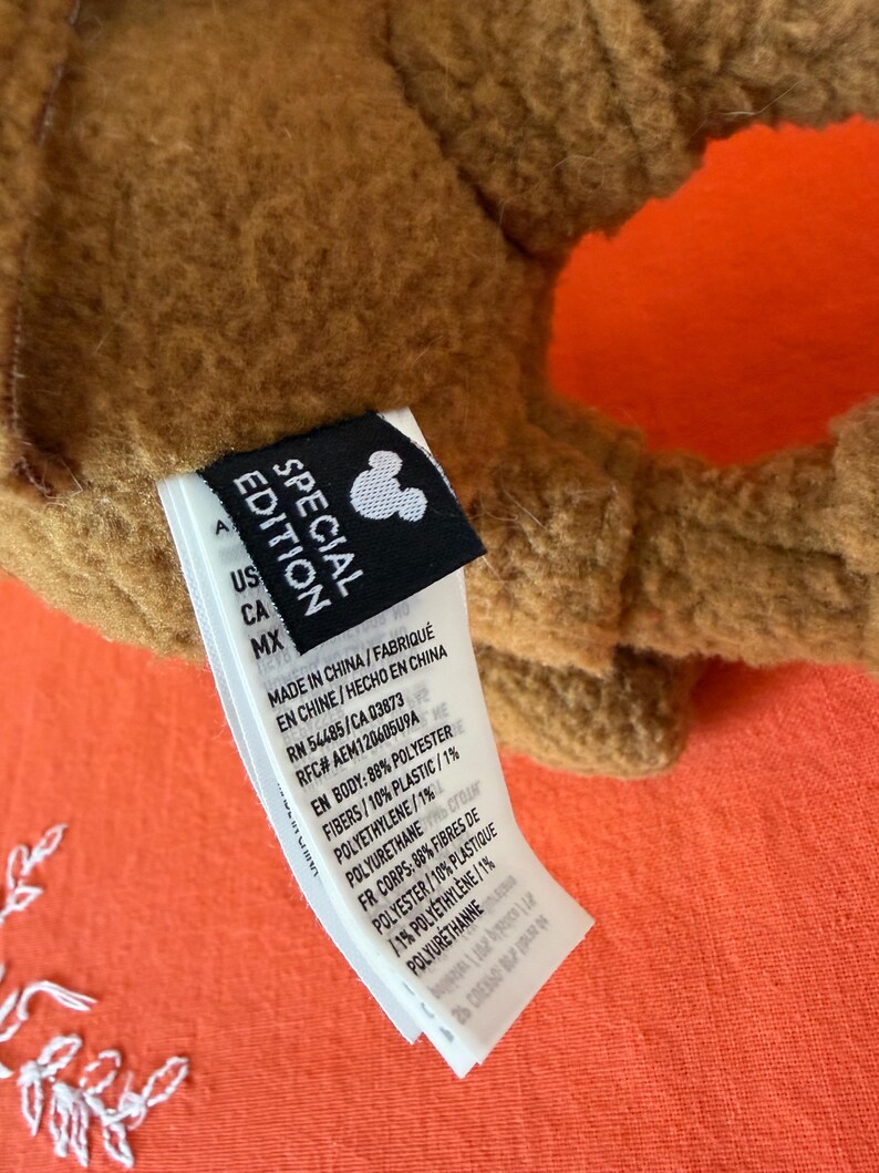 May include: Close-up of a brown plush toy with an orange accent. A black tag with the text "SPECIAL EDITION" and a Mickey Mouse silhouette is attached. The tag also includes manufacturing details.