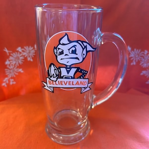 Peut inclure: Mug à bière en verre transparent avec une anse. Le mug présente un logo orange circulaire avec un personnage de dessin animé et le mot "BELIEVELAND" sur une bannière. Le mug est posé sur une surface orange.