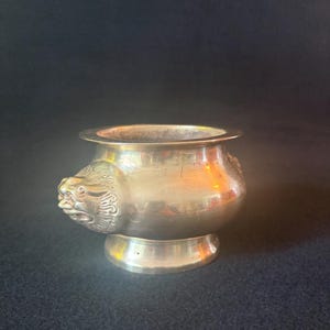 May include: A small, antique brass bowl with a detailed lion's head on one side. The bowl has a rounded body, a wide rim, and a pedestal base. The brass has a warm, golden hue, and the bowl is set against a dark background.